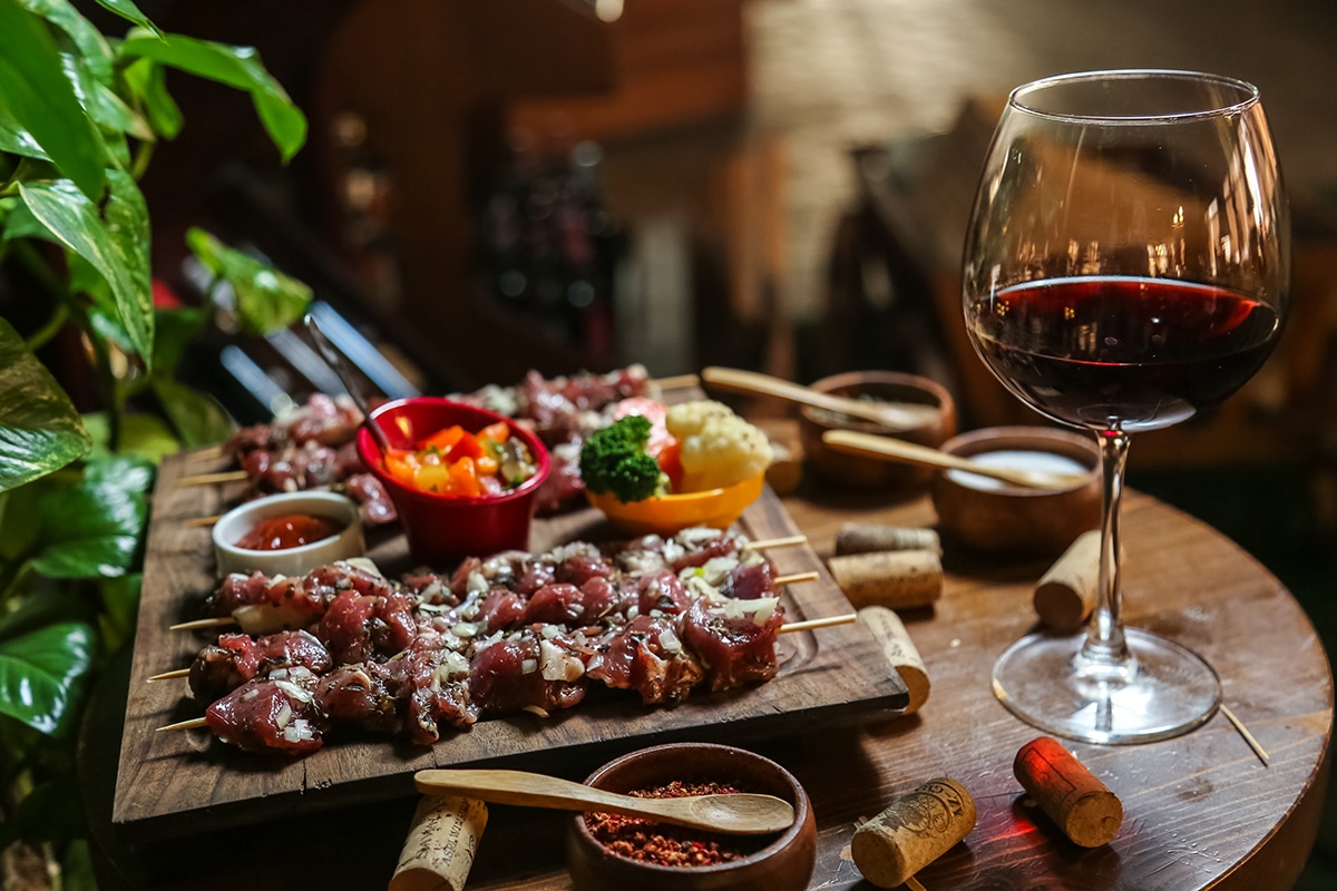 side view raw meat on skewers with vegetables and spices on a stand and a glass of red wine on the table