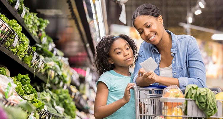 Mother and daughter grocery shopping in a supermarket, looking at a list while standing beside a cart near the produce section.