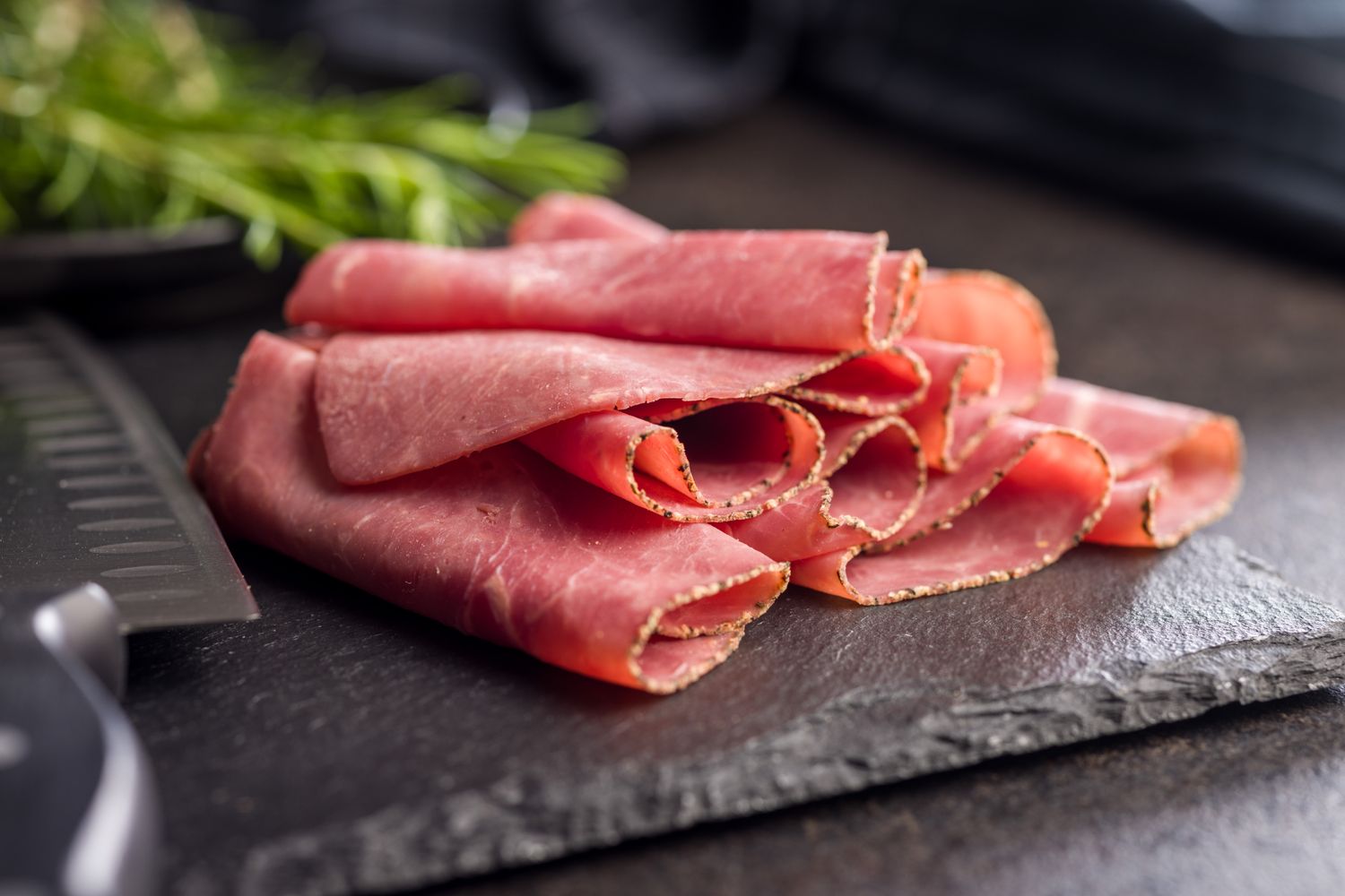 Thinly sliced roast beef rolled into curls on a dark slate board, with a knife and fresh herbs in the background.